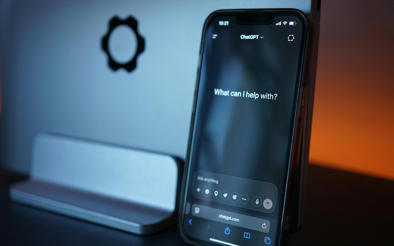ChatGPT interface open on a smartphone screen displaying the prompt "What can I help with?", placed against a dark background with a MacBook and ambient orange lighting